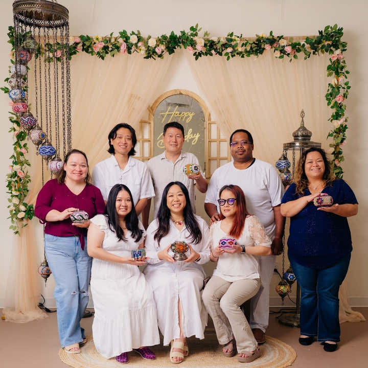 Group of eight people posing together holding painted decorative bowls in front of floral backdrop and hanging decor