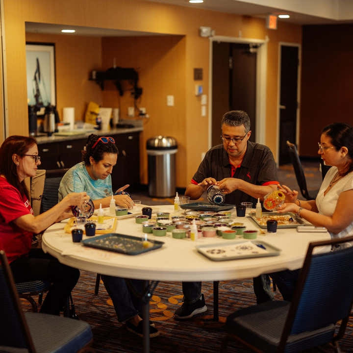 Four people sitting around a white table working on electronics and circuit boards in a modern workshop space with orange walls